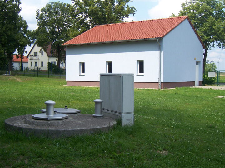 Das Bild zeigt das Wasserwerk in Buckow: im Vordergrund die Oberflächenaufbauten eines Brunnens im Hintergrund ein gepflegtes kleines, einstöckiges Haus.